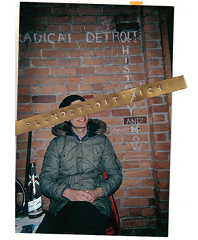 Picture of person in a green coat sitting in front of a brink wall that says 'Radical Detroit.' There is a sticker covering the person's face.
