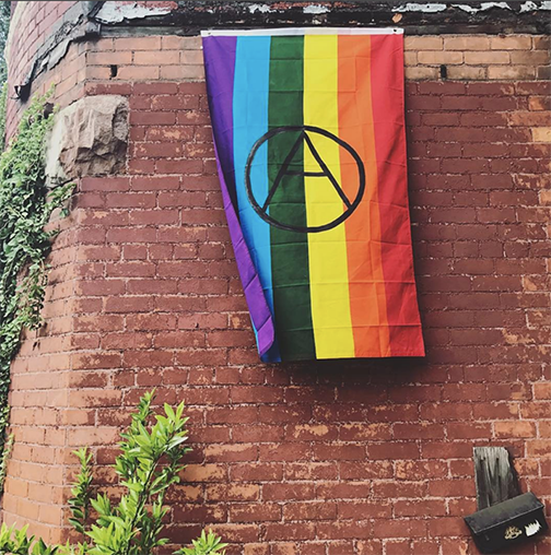 picture of a red brick wall with a rainbow pride flag with an anarchy sign painted on it in black hanging on it